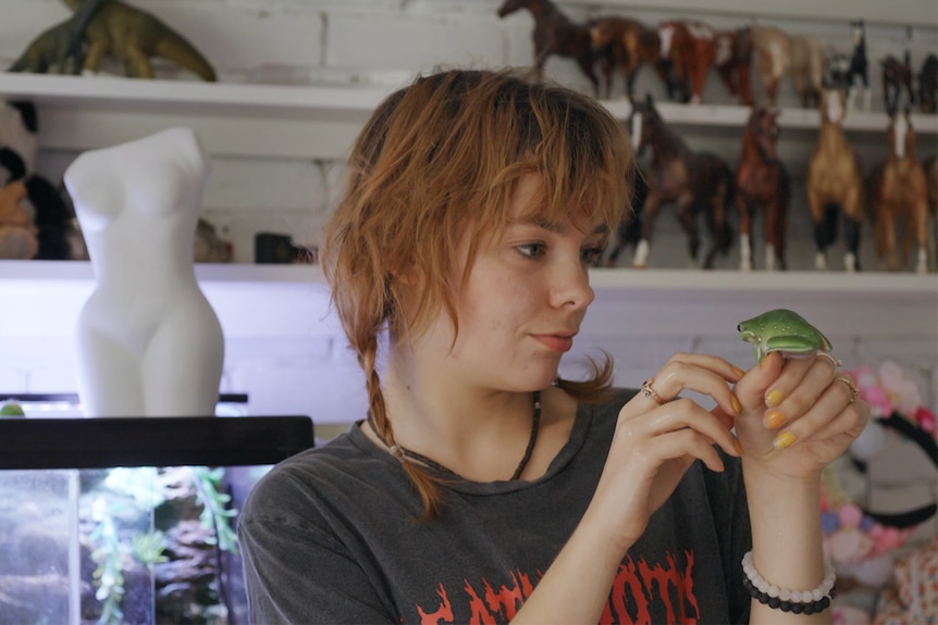 A young woman holds a frog up to her face, behind her are shelves of horse figurines.