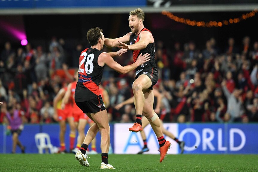 A male AFL player jumps in the air as he celebrates kicking a goal with a teammate.