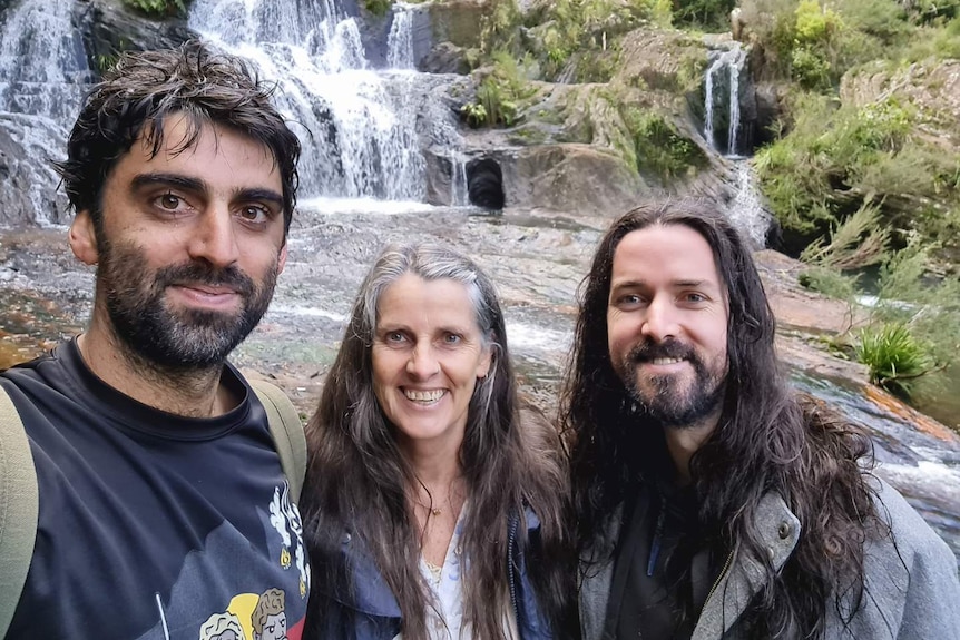 Two men and a woman stand together in front of a waterfall, smiling at the camera.