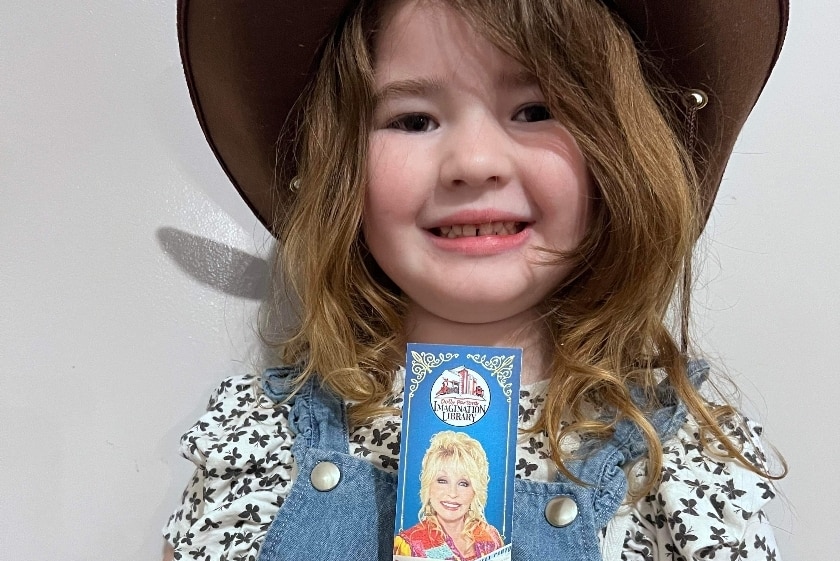 A young girl in a cowboy hat, holding a bookmark featuring Dolly Parton.