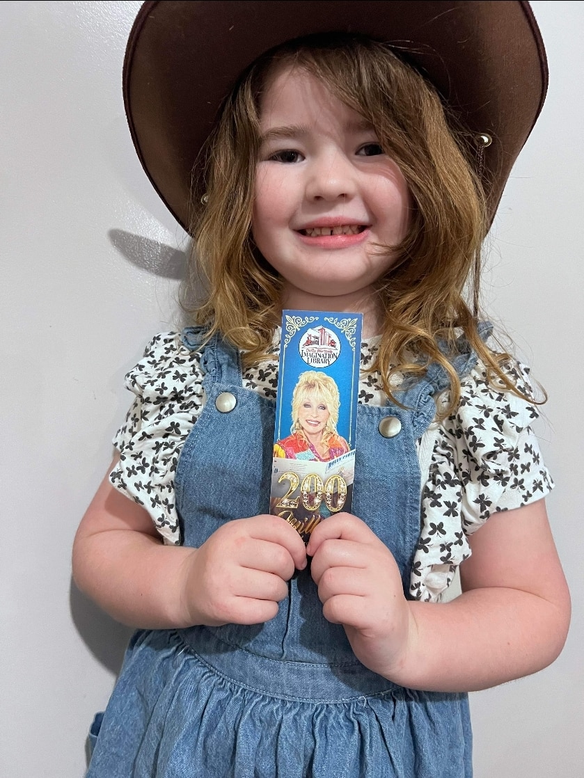 A young girl in a cowboy hat, holding a bookmark featuring Dolly Parton.