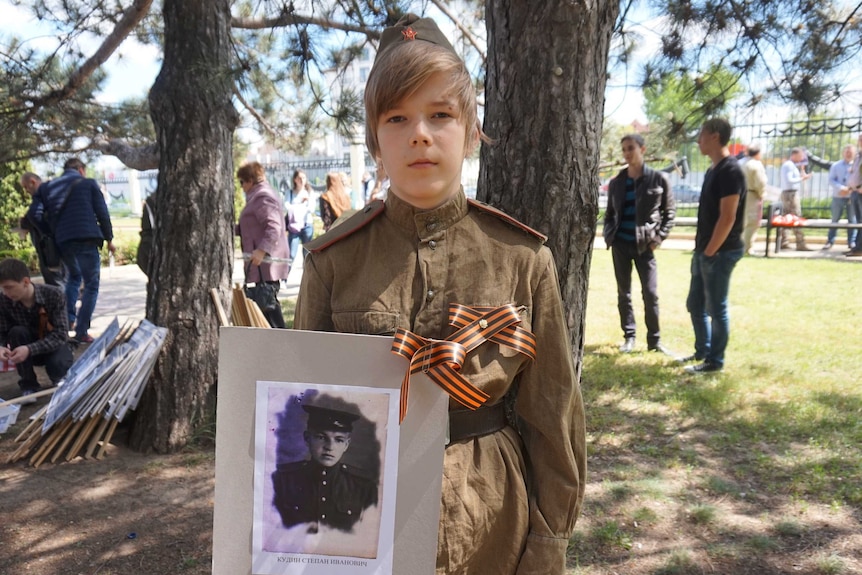 A young boy wearing a military uniform looks into the camera while holding an old photo of a soldier.