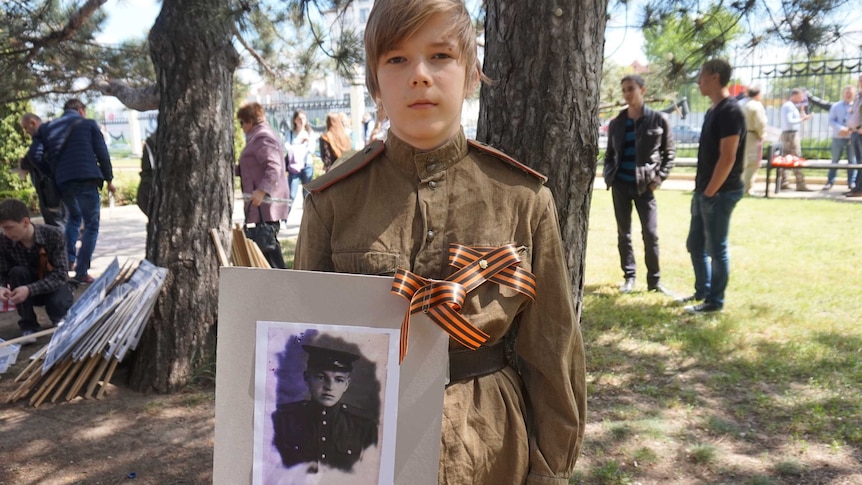 A young boy wearing a military uniform looks into the camera while holding an old photo of a soldier.