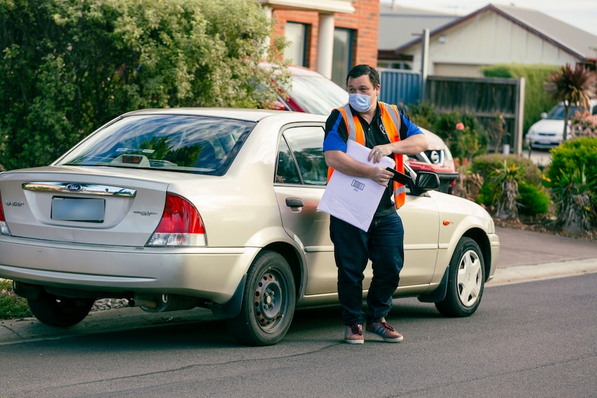 A man in an orange high-vis vest carries an envelope as he stands in front of a car on a suburban street. 