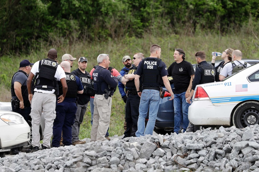 Police and people gather near a wooded area.