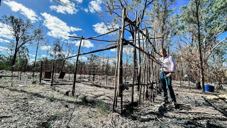 a man leans against a burnt-out fence next to a vegetable garden