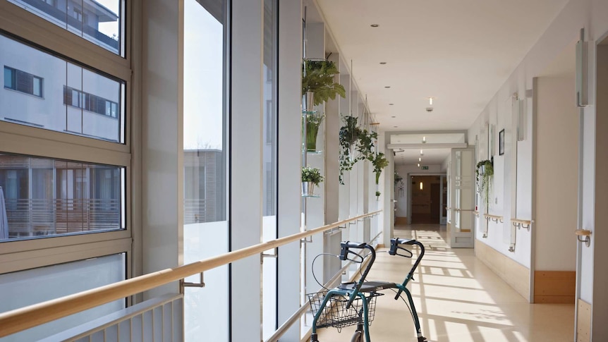 A walking frame sits in the foreground of a sunlit corridor in an aged care facility.