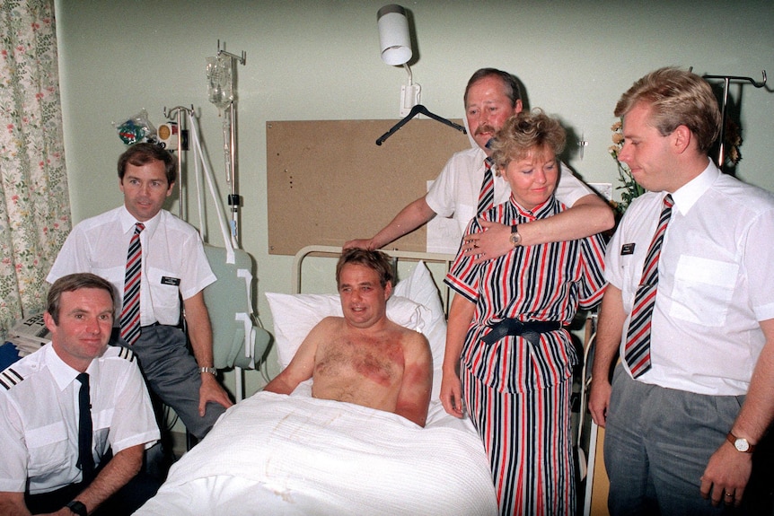 Four men and a woman in airline uniforms stand around a hospital bed where a shirtless man is sitting up