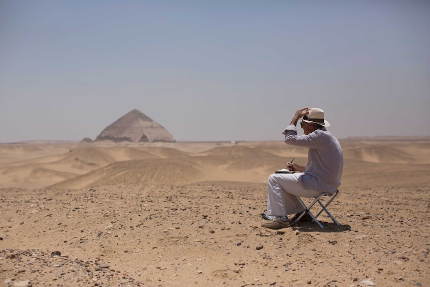 A member of an international delegation sits on a stool and sketches the site of the Bent Pyramid, seen in the distance.
