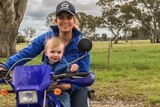 A woman wearing a blue workshirt matches her son sitting on a blue two wheel motorbike.