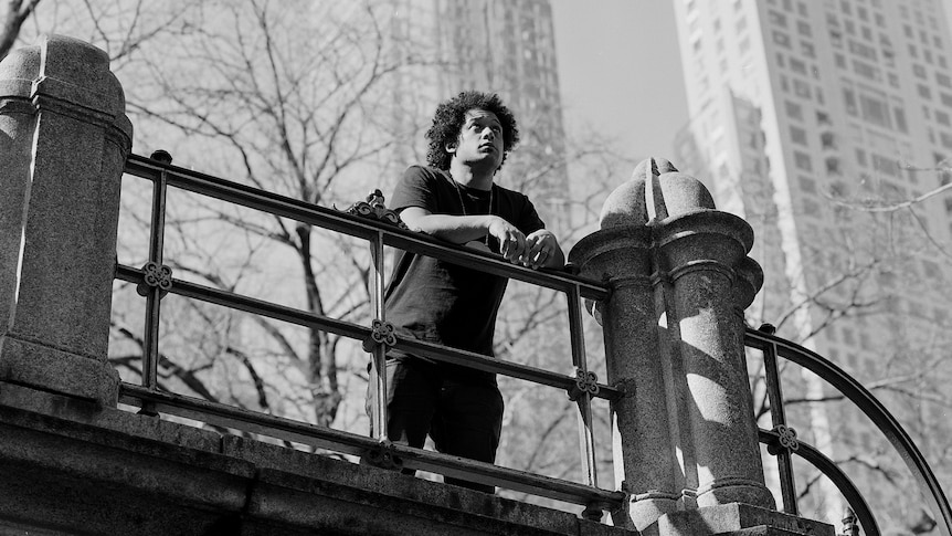 A black and white image of Makaya standing on a bridge looking out. Behind him are two large towers.