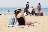 A woman sits reading a beach on the sand, dressed in swimmers and a hat.