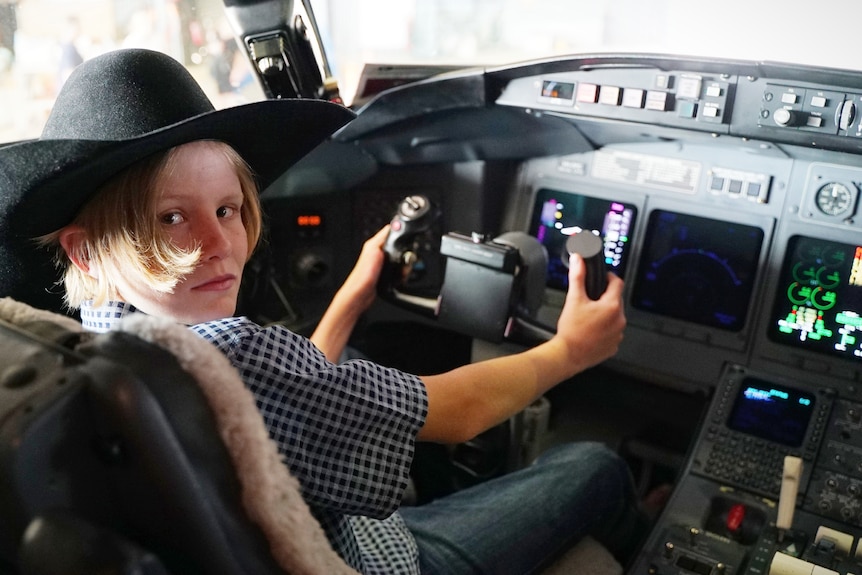 A boy in a cowboy hat sits in the cockpit of a plane with his hands on the controls.
