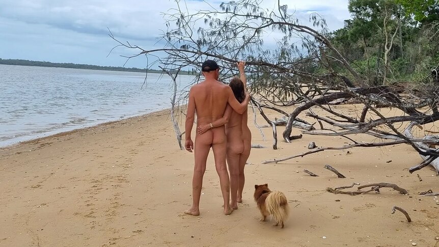 A naked man with his arm around a naked woman on a beach. A small dog stands behind them.