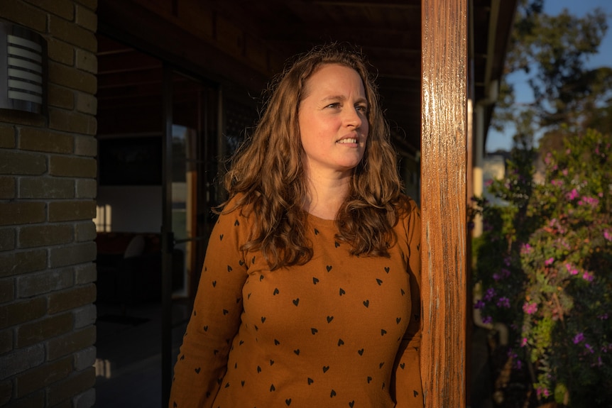 A woman with a tan jumper and long brown hair leans on a wooden pole on her front porch, looking out to the distance.