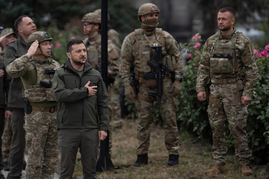 Un homme en robe kaki participe à une cérémonie de lever du drapeau national aux côtés de soldats en tenue de combat.