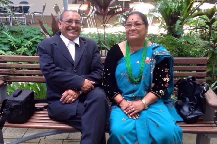 A man and a woman sit on a bench in front of some plants. She is wearing a sari.