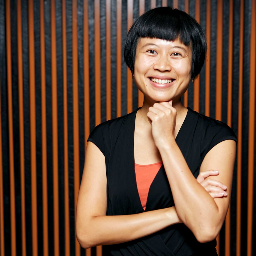 A woman with short black hair smiles with her hand near her chin and her arm crossed.