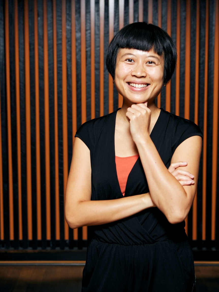 A woman with short black hair smiles with her hand near her chin and her arm crossed.