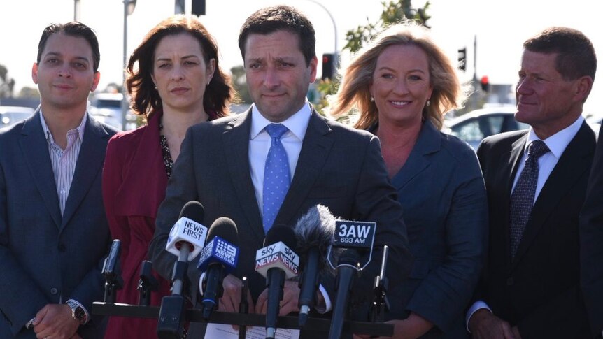 Matthew Guy speaks to reporters in front of a traffic intersection, flanked by his wife and three Liberal candidates.