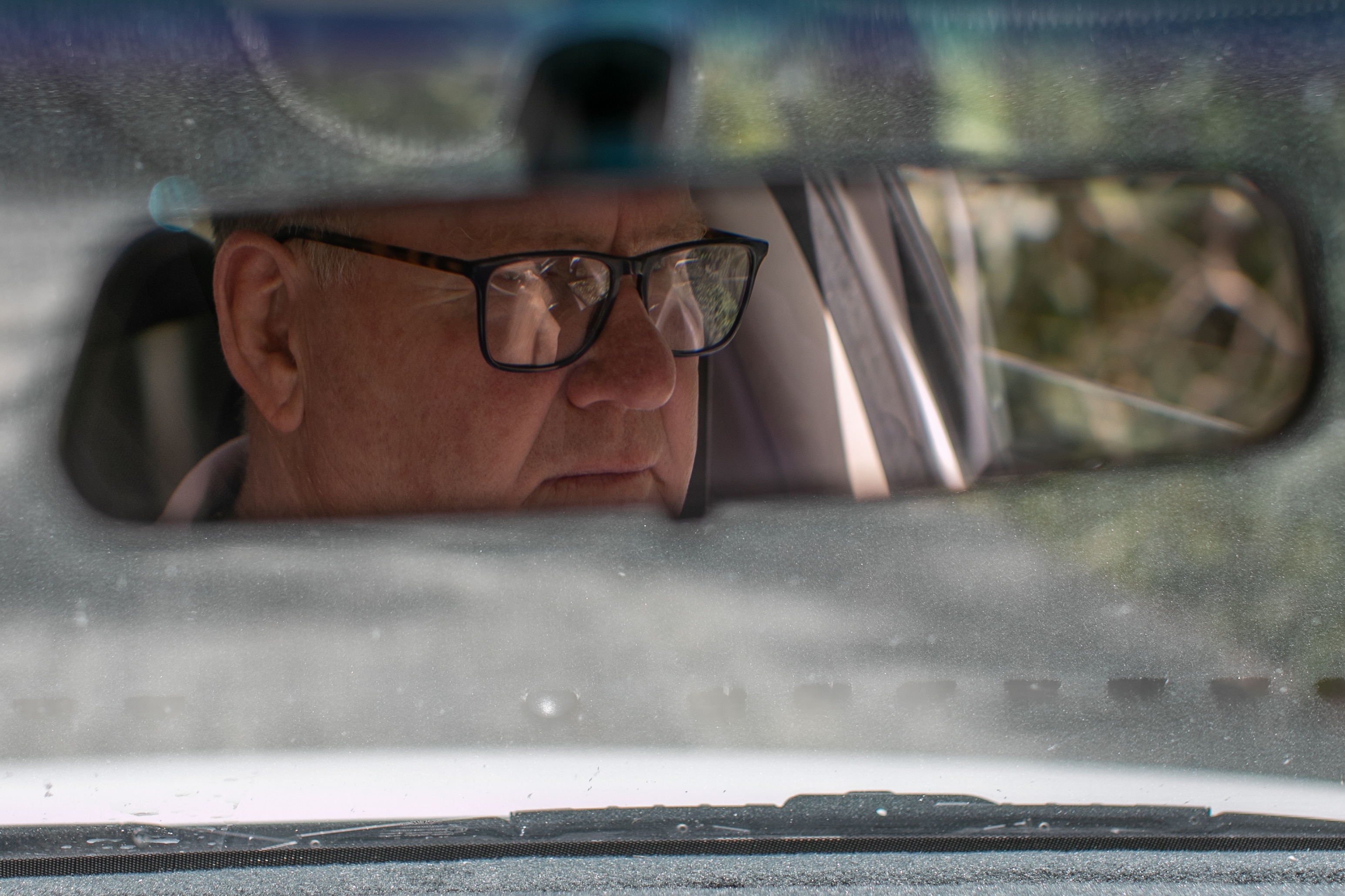 A man's face is seen in rear-vision mirror inside a car, he wears glasses and watches the road carefully