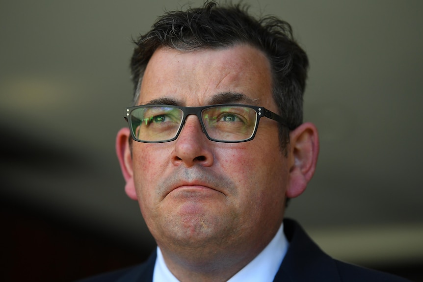 Daniel Andrews stands at a microphone outdoors in a shirt, jacket and blue tie wearing glasses.