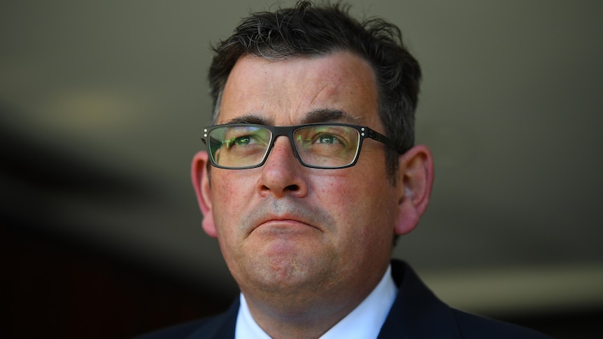 Daniel Andrews stands at a microphone outdoors in a shirt, jacket and blue tie wearing glasses.