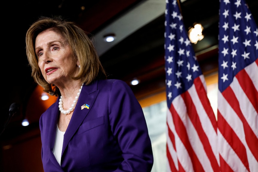 Woman with short hair in purple blazer with two US flags behind her.