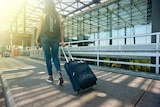 A woman holding a suitcase on wheels walks away from the camera.