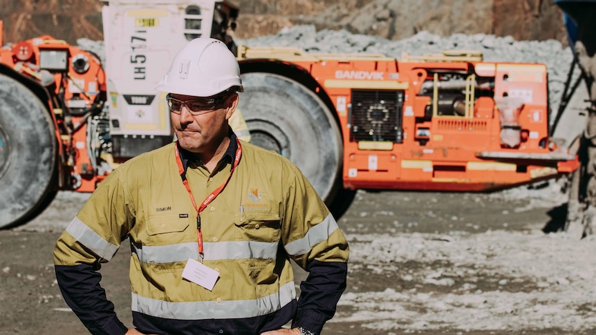 A man wearing high-vis workwear and a hard hat standing in front of mining machinery in a gold mine.  