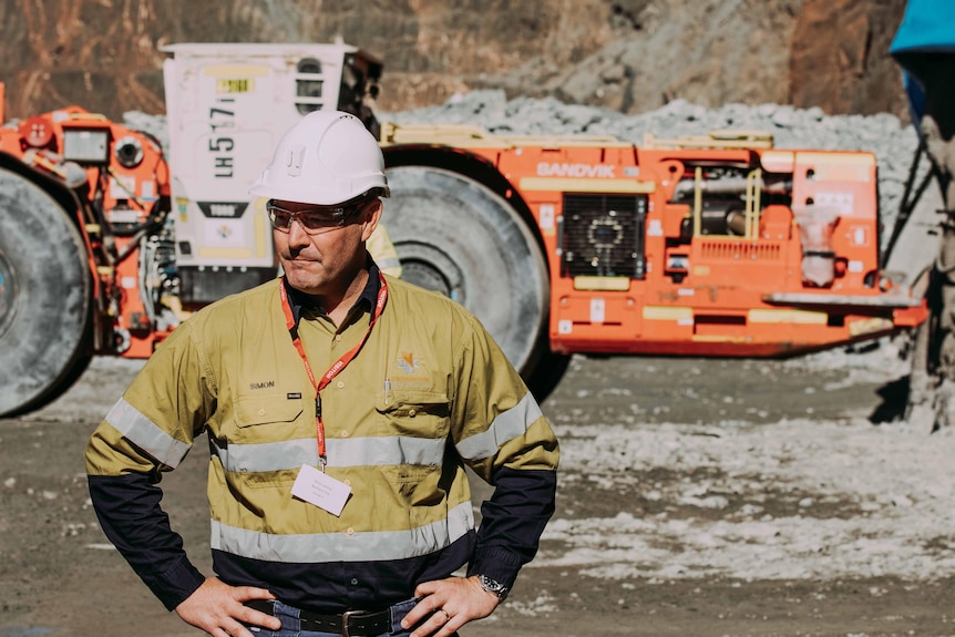 A man wearing high-vis workwear and a hard hat standing in front of mining machinery in a gold mine.  