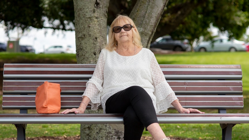 An older woman with blonde hair and wearing sunglasses and a white shirt sits outside on a park bench.