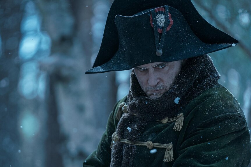 Joaquin Phoenix as Napoleon has a brooding expression and wears the iconic hat while snow falls in front of his face.