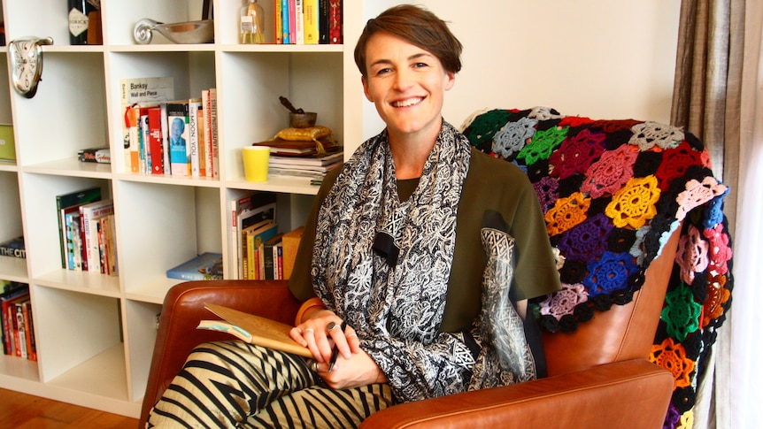 A woman sits in a leather armchair, holding a notebook and smiling at the camera.