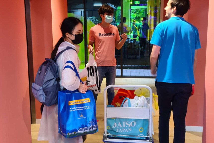 Two students stand near a lift wearing masks as a man helps them push a trolley with moving bags on it.