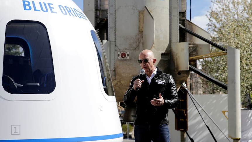 A short man in black leather jacket speaks into microphone outside on sunny day beside blue origin aircraft