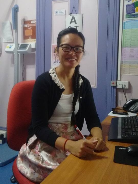 A female doctor with dark hair and glasses sitting in front of a computer in a consultation room.