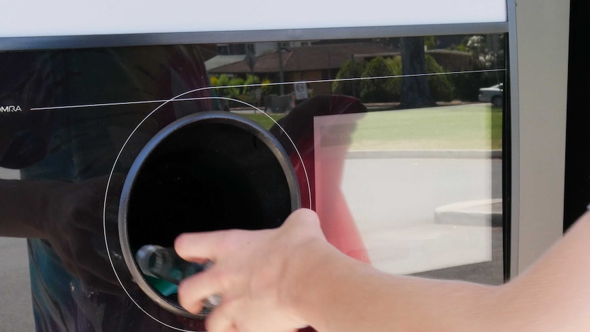 A close up of a hand putting a glass bottle into a return and earn chute.