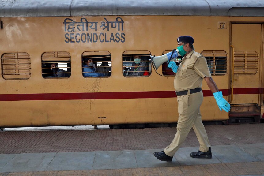 A police officer in a face mask and gloves speaks into a megaphone while walking past a train