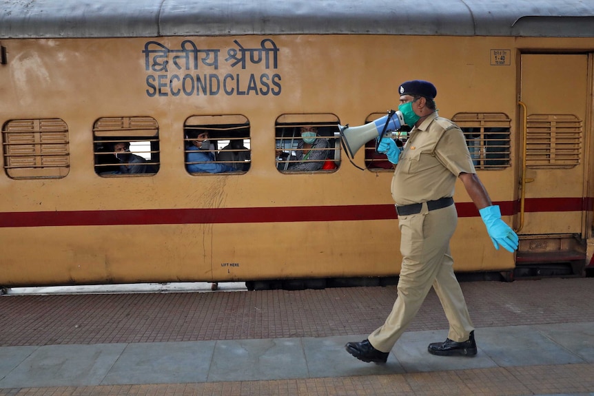 A police officer in a face mask and gloves speaks into a megaphone while walking past a train