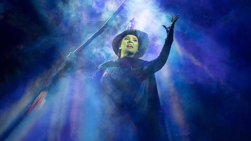 Sheridan Adams as Elphaba. She is holding a broom and surrounded by mist.