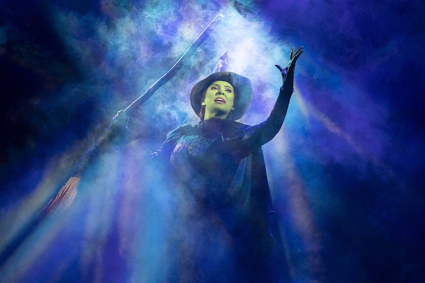 Sheridan Adams as Elphaba. She is holding a broom and surrounded by mist.