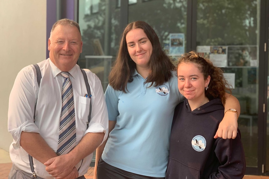 An older male (the school principal) standing next to two female year 11 students. They are all smiling.