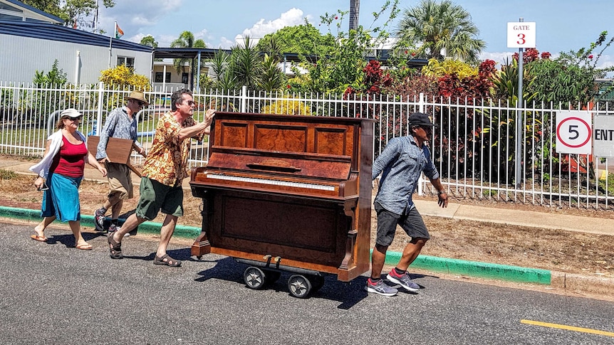 Comedic scene of four people escorting a wooden upright piano on wheels down a suburban street.