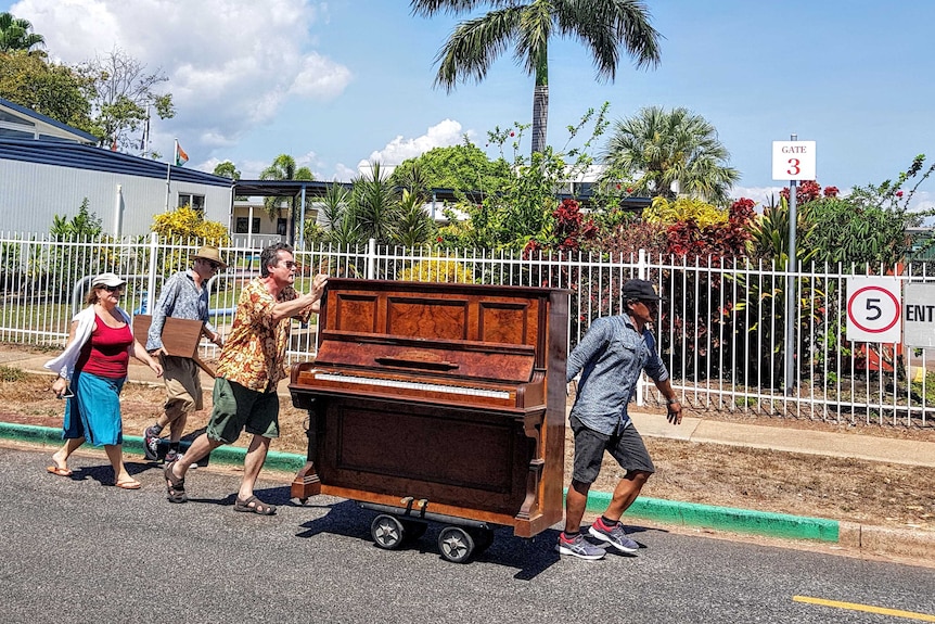 Comedic scene of four people escorting a wooden upright piano on wheels down a suburban street.