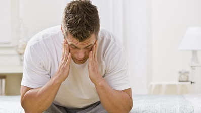 A man is seated on a bed looking frustrated.