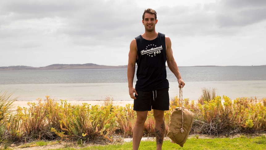 A muscular man in black tank top and shorts stands holding a burlap sack.