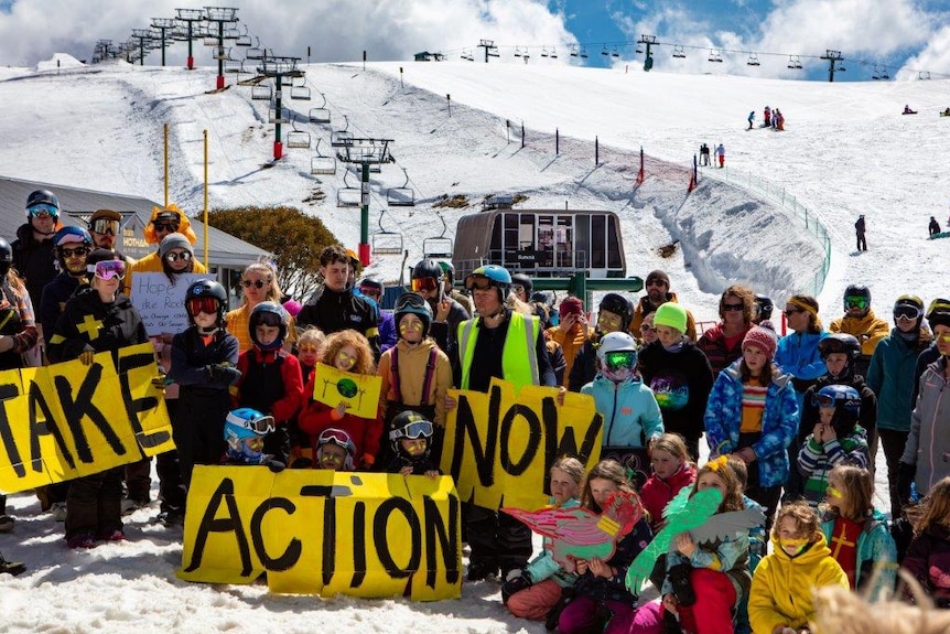 A group of protesters gathered in the snow holding placards with a ski lift behind them.