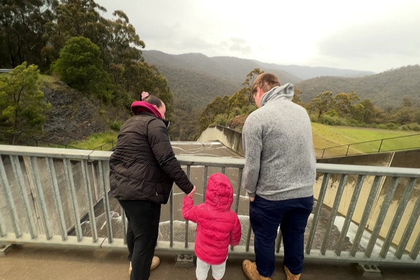 Three people standing on a bridge watching water flow out of a full dam.