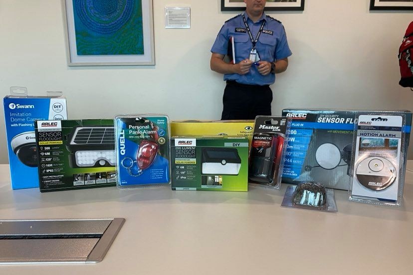 A number of security devices, in packaging, sit on a table. A police officer stands behind.